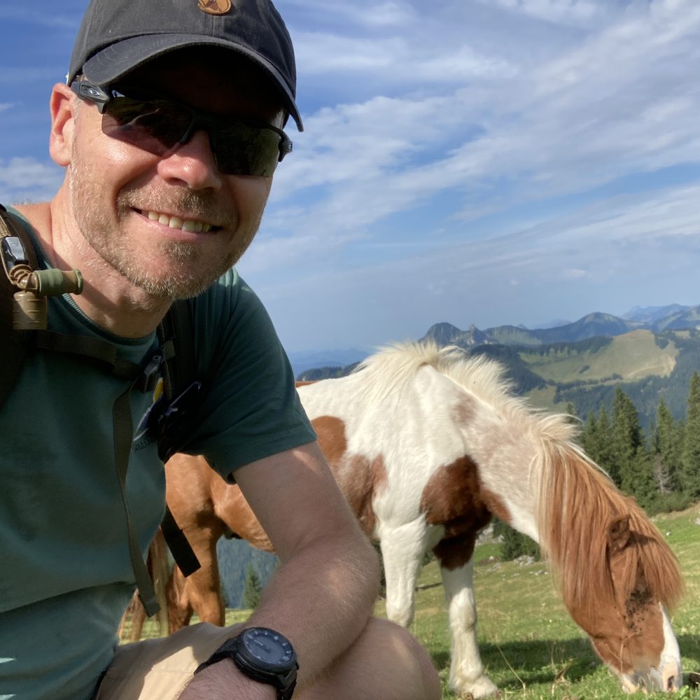 Alexander Siering mit Kappe und Sonnenbrille, daneben ein grasendes Pony auf einer Bergwiese